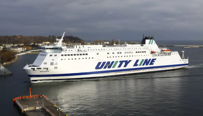 Biały prom Unity Line "Polonia" płynie po wodzie, w tle widać port i zachmurzone niebo.