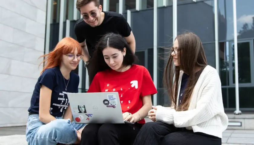 Cztery osoby siedzą na schodach przed budynkiem, patrząc na laptopa.