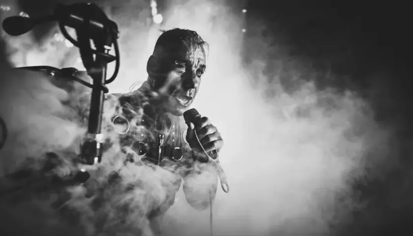 Lindemann w dymie, z mikrofonem w dłoni, w mrocznym klimacie. Jego twarz jest intensywna, jakby śpiewał "Knebel".