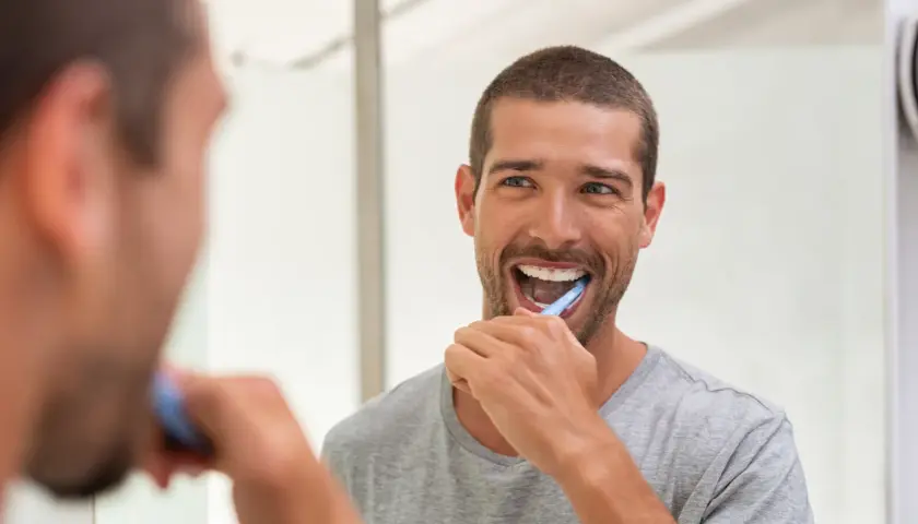 Uomo sorridente si lava i denti davanti allo specchio, un gesto quotidiano per la prevenzione odontoiatrica.