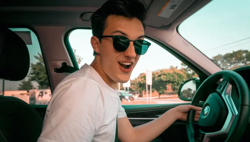 Joven con gafas de sol sonríe emocionado al volante de su coche novel, listo para una aventura.