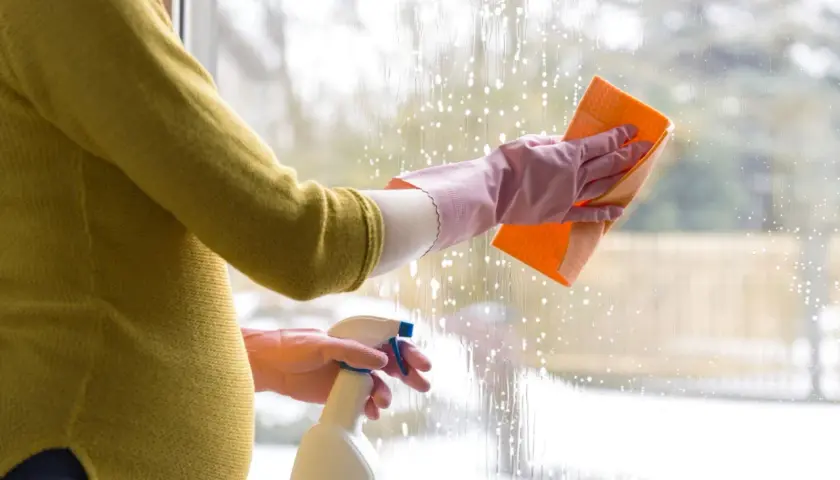 Ciężarna kobieta w rękawiczkach myje okno gąbką i płynem w sprayu.