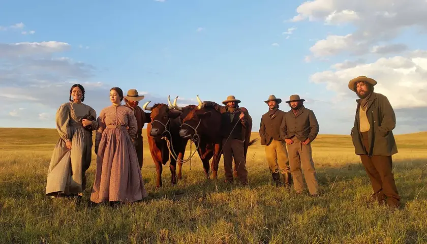 Un gruppo di persone in abiti d'epoca, con due buoi, in un campo dorato. Scena da "La ballata di Buster Scruggs", un film che merita una recensione.