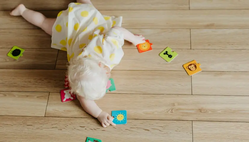 Dziecko z jasnymi włosami, w żółtej sukience w kropki, bawi się na podłodze. Rozrzucone są puzzle ze zwierzątkami i słońcem. To moment, kiedy dziecko zaczyna się obracać.