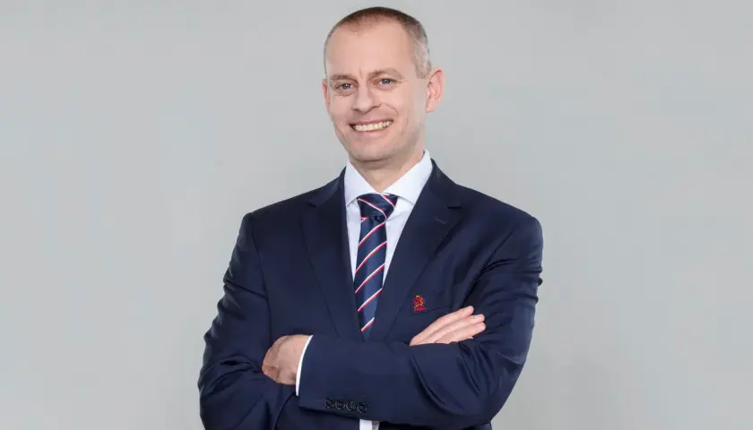 Marcin Włodarski, trener, w granatowym garniturze i krawacie, z rękami skrzyżowanymi na piersi, uśmiecha się do obiektywu.