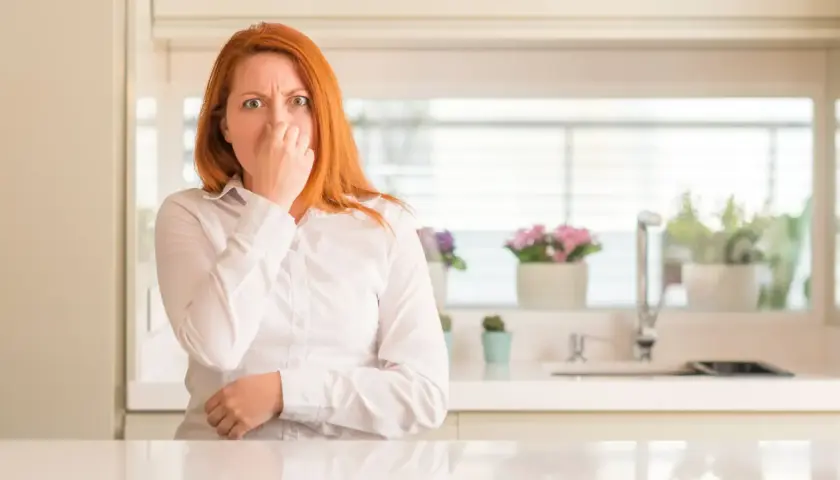 Mujer pelirroja se tapa la nariz, con cara de asco, por unos gases con olor a huevos podridos en la cocina.