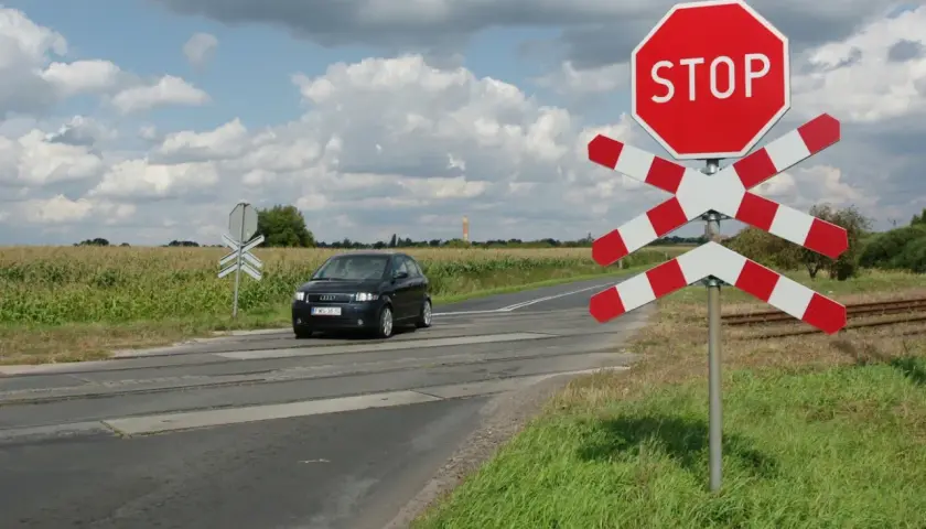 Czarny samochód zbliża się do przejazdu kolejowego z czerwonym znakiem STOP i białoczerwonym krzyżem.