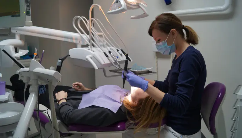 Dentystka w masce i rękawiczkach pracuje przy pacjencie. W gabinecie widoczny sprzęt do stomatologii.
