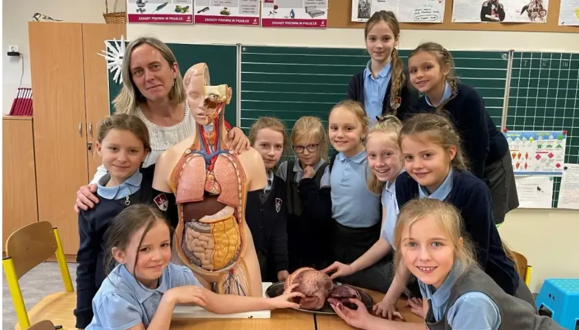 Nauczycielka i uczennice przy modelu anatomicznym. Jedna z dziewczynek trzyma serce na dłoni, jakby ofiarowywała je w geście nauki.