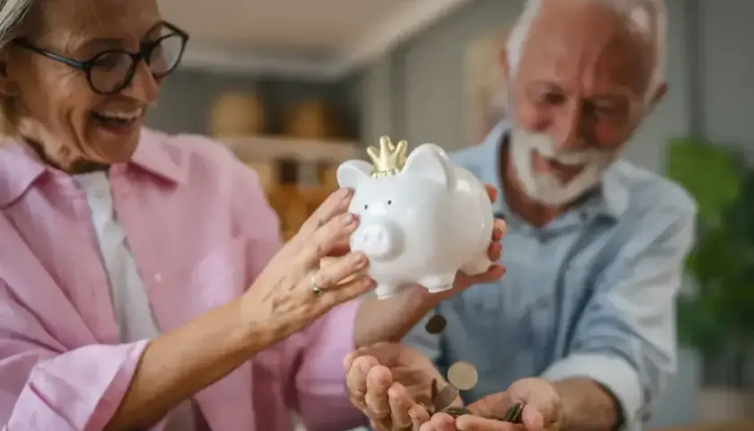 Uśmiechnięta para seniorów wsypuje monety do skarbonki-świnki. Czekają na moment, kiedy wyrównają emerytury czerwcowe.