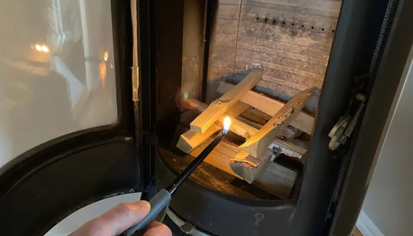 En hand tänder ved i en kamin. Det syns lite tjära i skorstenen.
