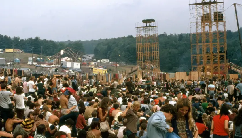 Tłum ludzi na festiwalu muzycznym, gdzie jest Woodstock. Widać scenę, wieże głośnikowe i namioty.