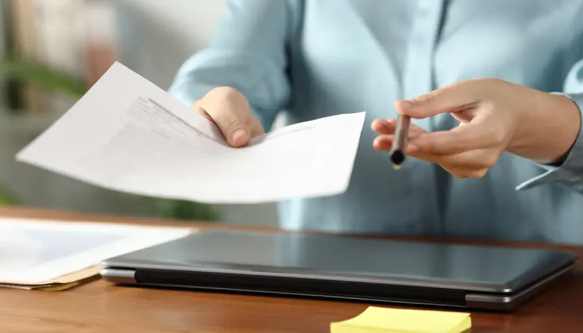 Dłoń podaje dokument, sugerując omawianie różnych form zatrudnienia. Na biurku leży laptop i żółta karteczka.