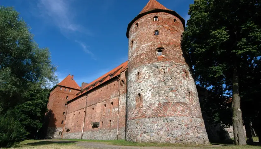 Zamek w Bytowie, potężna ceglana twierdza z kamienną podstawą wieży, otoczona zielenią pod błękitnym niebem.
