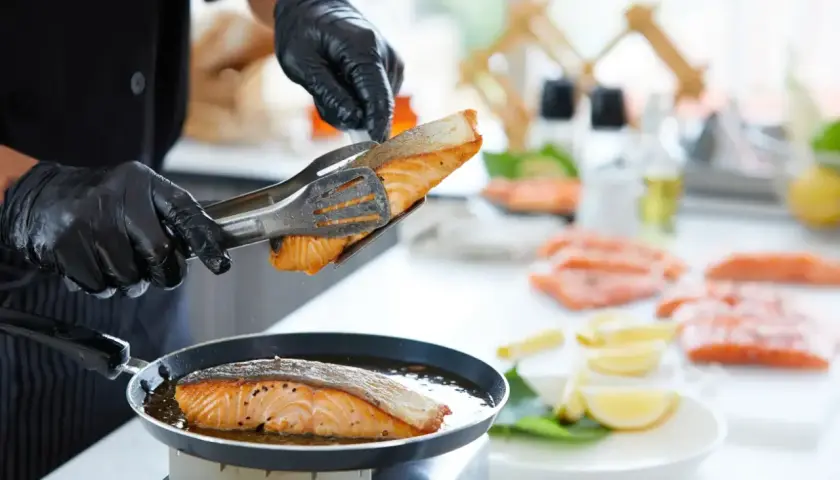 Szef kuchni w czarnych rękawiczkach kładzie łososia na patelni. Smażenie ryby na złoto to właśnie sauté.