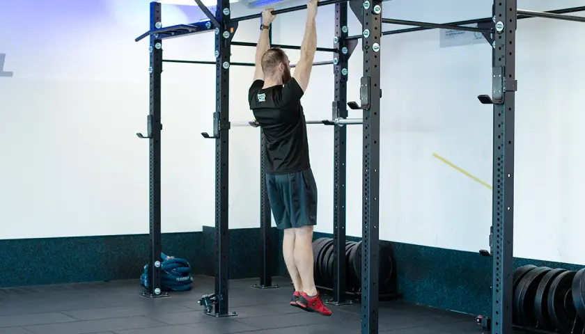 Mężczyzna wykonuje scapular pull ups na drążku. Ćwiczy plecy i ramiona w siłowni.