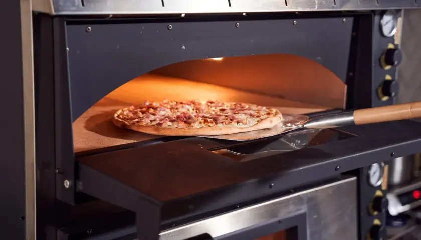 Une pizza garnie est enfournée dans un four à pizza professionnel. La pelle à pizza glisse sous la pâte dorée, prête à être cuite.