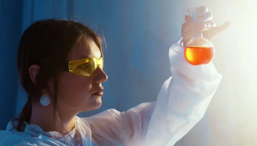 Naukowczyni w okularach ochronnych analizuje płyn w kolbie. To jej praca w dziedzinie biotechnologii.