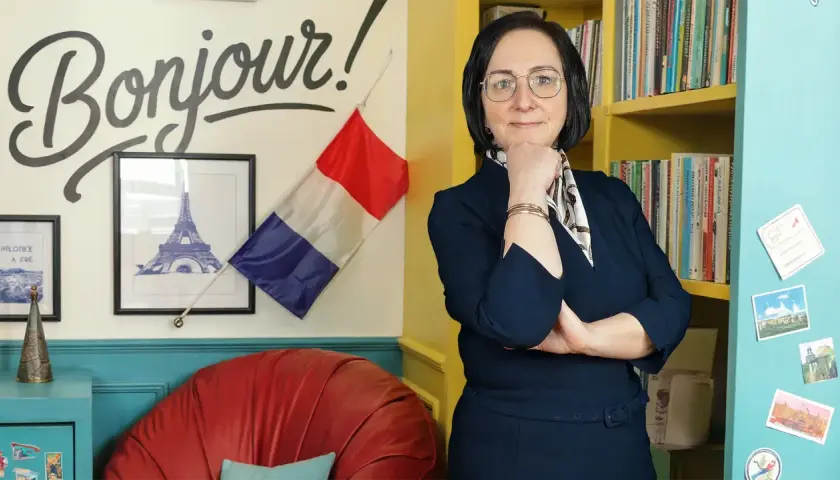 Kobieta w okularach stoi przed ścianą z napisem "Bonjour!", francuską flagą i wieżą Eiffla.