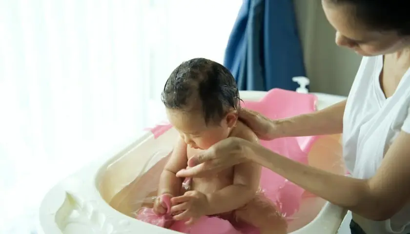 Mama delikatnie kąpie dziecko w wanience. Woda jest ciepła, a maluch bawi się zabawką. Może dodano krochmal do kąpieli dla miękkości skóry?