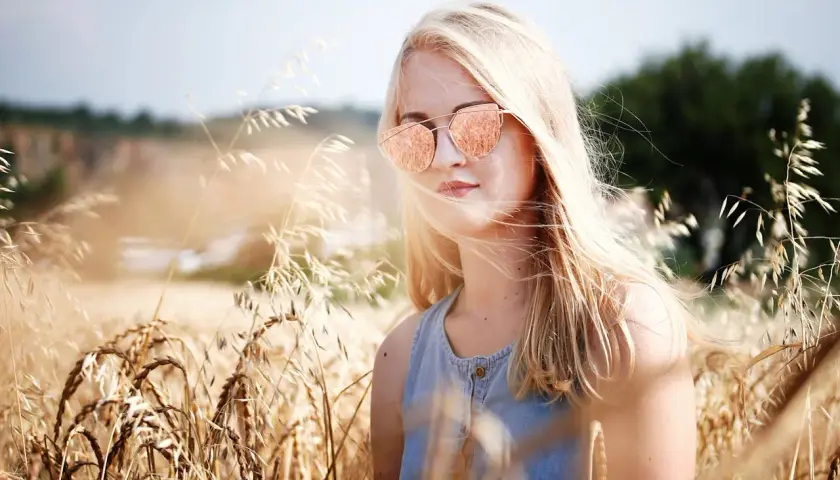 Dziewczyna z piaskowym blond włosami w okularach przeciwsłonecznych stoi w złotym polu pszenicy.