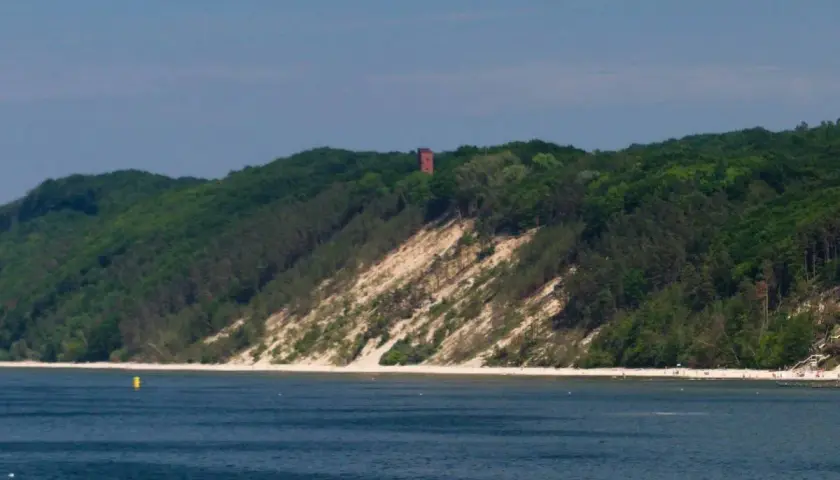 Zielone wzgórza porośnięte lasem schodzą ku morzu, odsłaniając piaszczystą, białą górę. Na szczycie majaczy ceglana wieża.