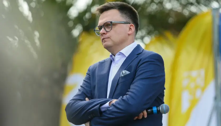 Szymon Hołownia w garniturze i okularach, z mikrofonem w ręku, na tle żółtych flag.