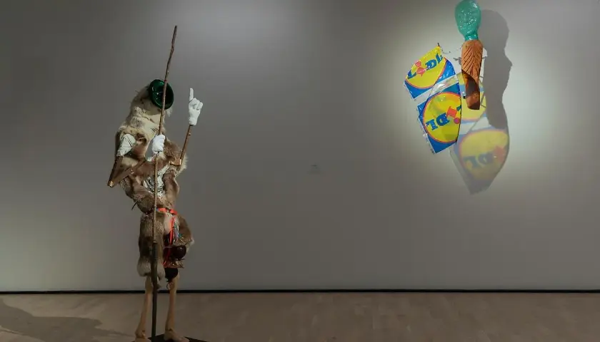 Konsthallar Sverige: En surrealistisk figur med päls och en pinne står bredvid en väggskulptur med Lidl-påsar och ett grönt huvud.