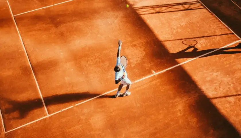 Agassi tenisista w akcji, serwuje na ceglanym korcie. Cień rakiety i sylwetka gracza rzucają się na pomarańczową nawierzchnię.