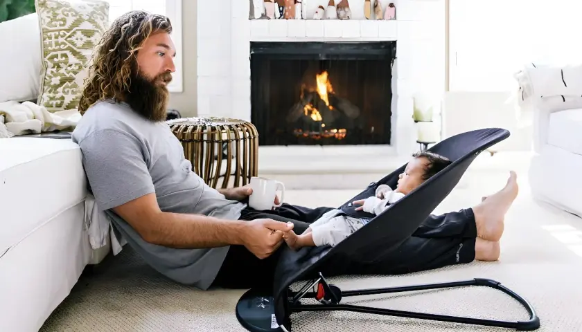 En man med skägg och en bebis i en babysitter. Elden sprakar i öppna spisen. Denna babysitter bäst i test gör pappan glad.