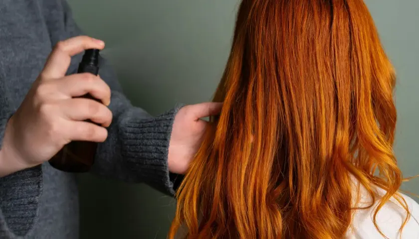 Persona rocía un producto en el cabello fino y quebradizo de color naranja rojizo para darle brillo y protección.