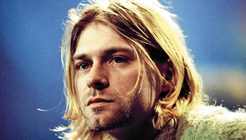 Kurt Cobain, ikona zespołu Nirvana, z charakterystycznymi blond włosami i niebieskimi oczami, na tle niebieskiego tła.