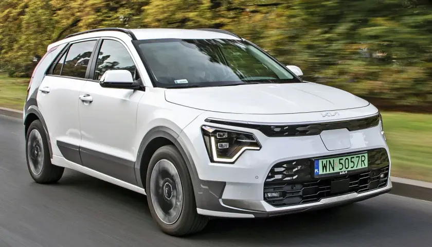 Biały Kia e-Niro jedzie po drodze, otoczony zielenią drzew.