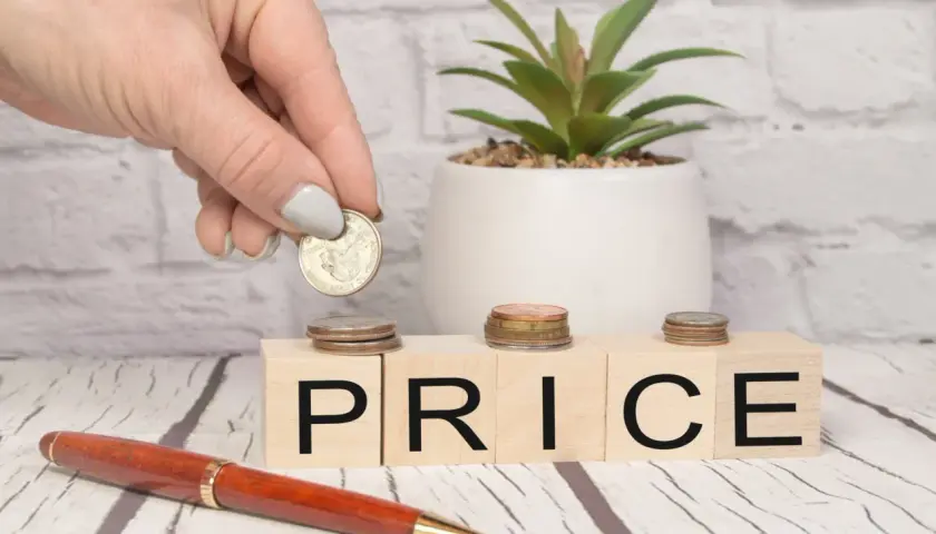 Dłoń kładzie monetę na kostce z literą "P", obok kostek z napisem "RICE". Zastanawiasz się, o ile służebność obniża wartość nieruchomości?