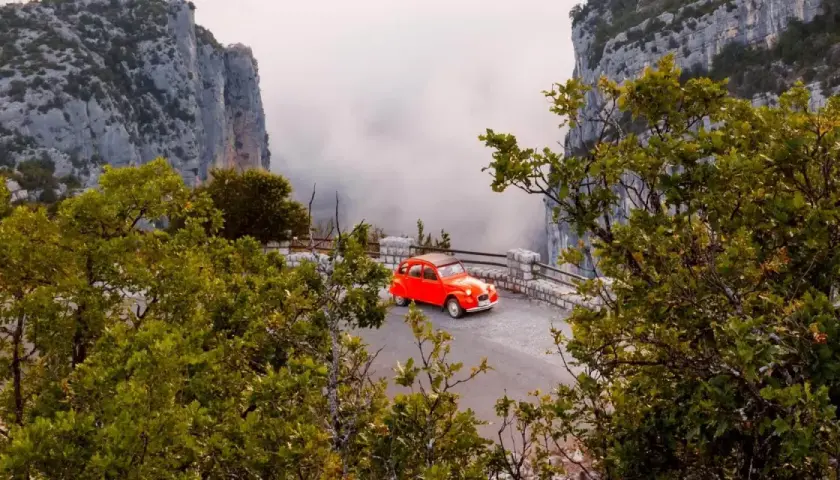 Une petite voiture rouge se gare sur la route des Gorges du Verdon, offrant une vue spectaculaire sur les falaises et la brume.