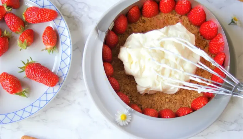 Préparation d'un cheesecake fraise : crème fouettée sur une base de biscuits et fraises fraîches.