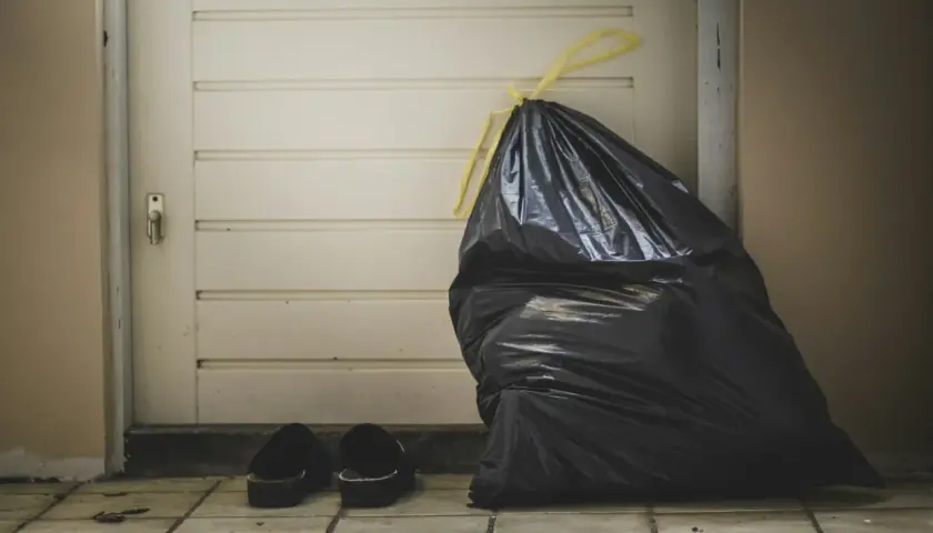 A full black trash bag and a pair of slippers sit by a closed door, symbolizing the need for lean budgeting to discard unnecessary expenses.