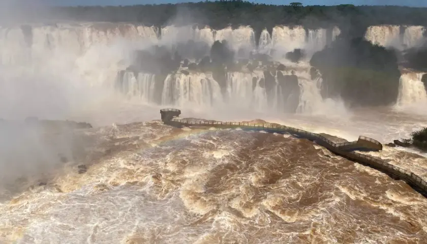 Mächtige Iguazú-Wasserfälle stürzen in schlammbraunes Wasser. Ein Steg führt durch den Sprühnebel, ein Regenbogen funkelt.