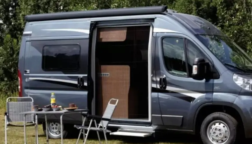 Un camping-car gris avec une moustiquaire plissée de porte ouverte. Une table et des chaises sont installées dehors.