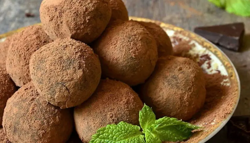 Un mucchio di tartufi al cioccolato facili e veloci, spolverati di cacao, su un piatto decorato, con menta fresca e un pezzetto di cioccolato fondente.
