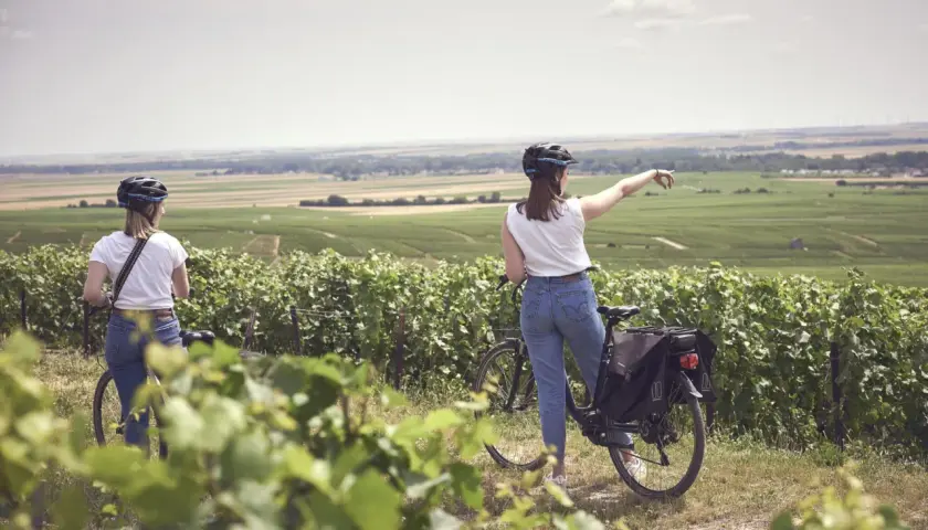 Two women on a champagne bike tour point towards rolling vineyards under a hazy sky.