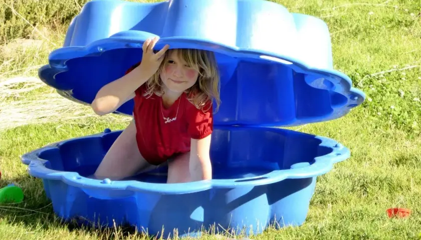Niña sonríe bajo una tapa azul de arenero, disfrutando de actividades de verano. ¡Diversión acuática para niños de 2 años!