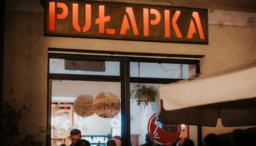 Neonowy napis "PUŁAPKA" nad wejściem do lokalu. W środku widać ludzi i dekoracje.