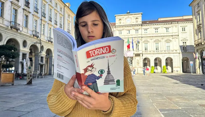 Bambina legge una guida "Torino con Viaggiapiccoli" in una piazza torinese. La libreria viaggi è perfetta per scoprire la città.