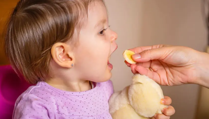 Małe dziecko z otwartymi ustami czeka na kawałek banana. Po biegunce u dziecka ważne jest nawadnianie i lekkie posiłki.