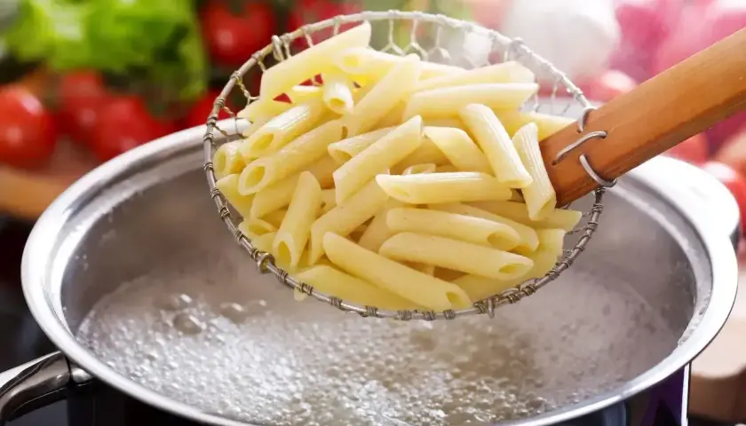 Penne makaron al dente w cedzaku nad garnkiem bulgoczącej wody. W tle pomidory i sałata.