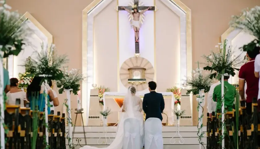 Le déroulement du mariage à l'église commence avec le couple devant l'autel, sous un crucifix. Les invités sont assis dans les bancs décorés de fleurs.