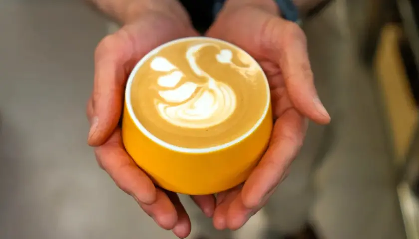 Dłonie trzymają żółtą filiżankę z kawą, na której artystycznie uformowano łabędzia. Zastanawiasz się, ile kofeiny ma espresso z ekspresu?