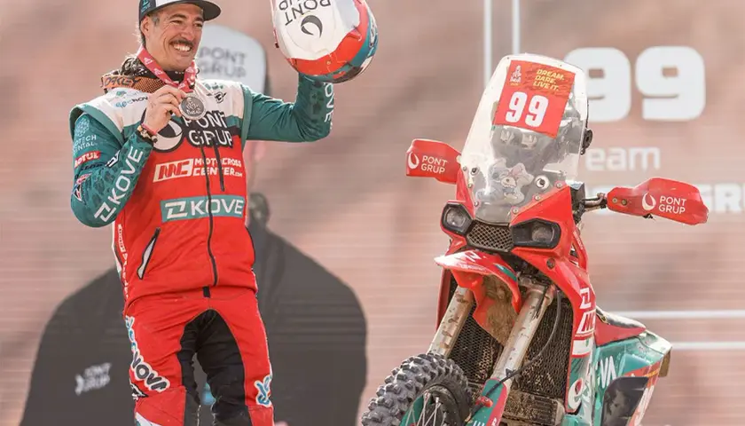 Piloto celebra victoria junto a su moto china Dakar. Casco y medalla en mano, luce orgulloso su equipación.