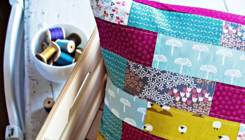 Buntes Patchwork-Kissen zeigt, wie man Stoffreste sinnvoll verwerten kann. Kleine Schafe und Blumenmuster zieren die Kissenhülle.